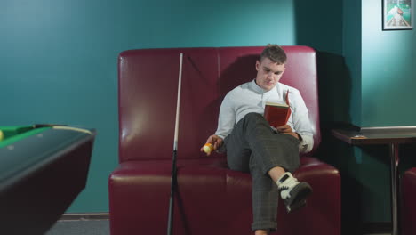 young man in white shirt and plaid pants sits on red sofa in billiard room, reading book while rolling orange billiard ball in hand. cue stick leans beside him. green pool table and dim lighting