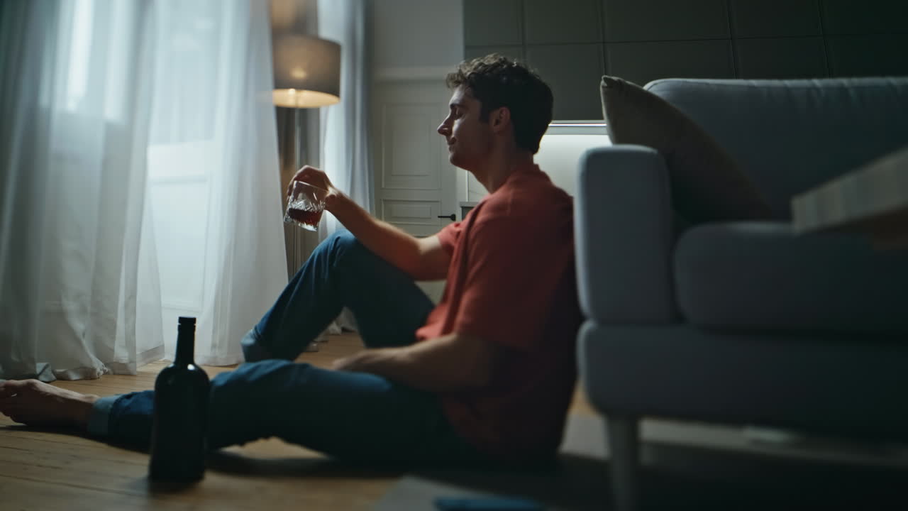 Alcohol addicted man suffering at empty night interior. Sad guy drinking bourbon