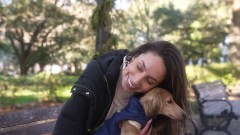 una mujer joven besa afectuosamente a su perro dachshund weiner en un banco del parque afuera en una tarde de otoño, amistad, sonrisa, dueño, acurrucarse, vincularse, amor, cachorro, regazo, abrazo, dulzura