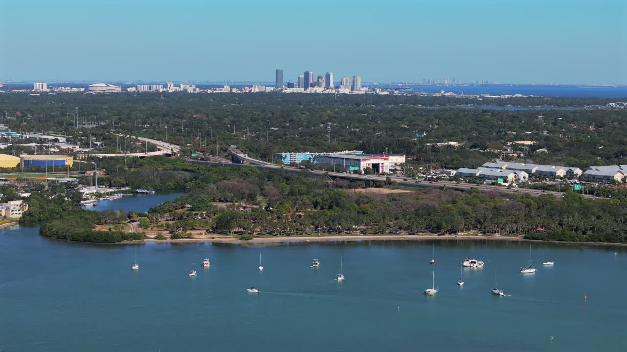 Aerial panning view of Maximo Park's natural shoreline with anchored sailboats and yachts in the bay, framed by highway ramp to Downtown Saint Petersburg skyline in Florida