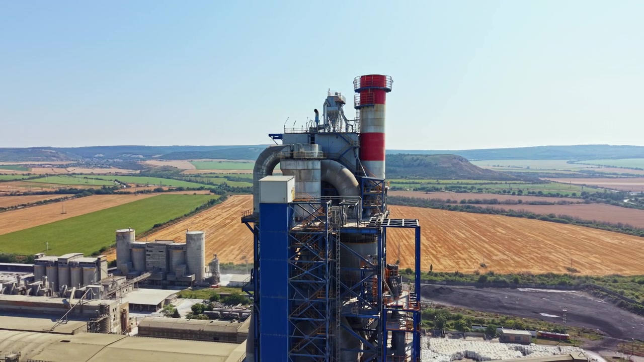 Alto edificio industriale con ciminiera visto dall'alto, circondato da campi e colline in una zona rurale durante una giornata limpida.
