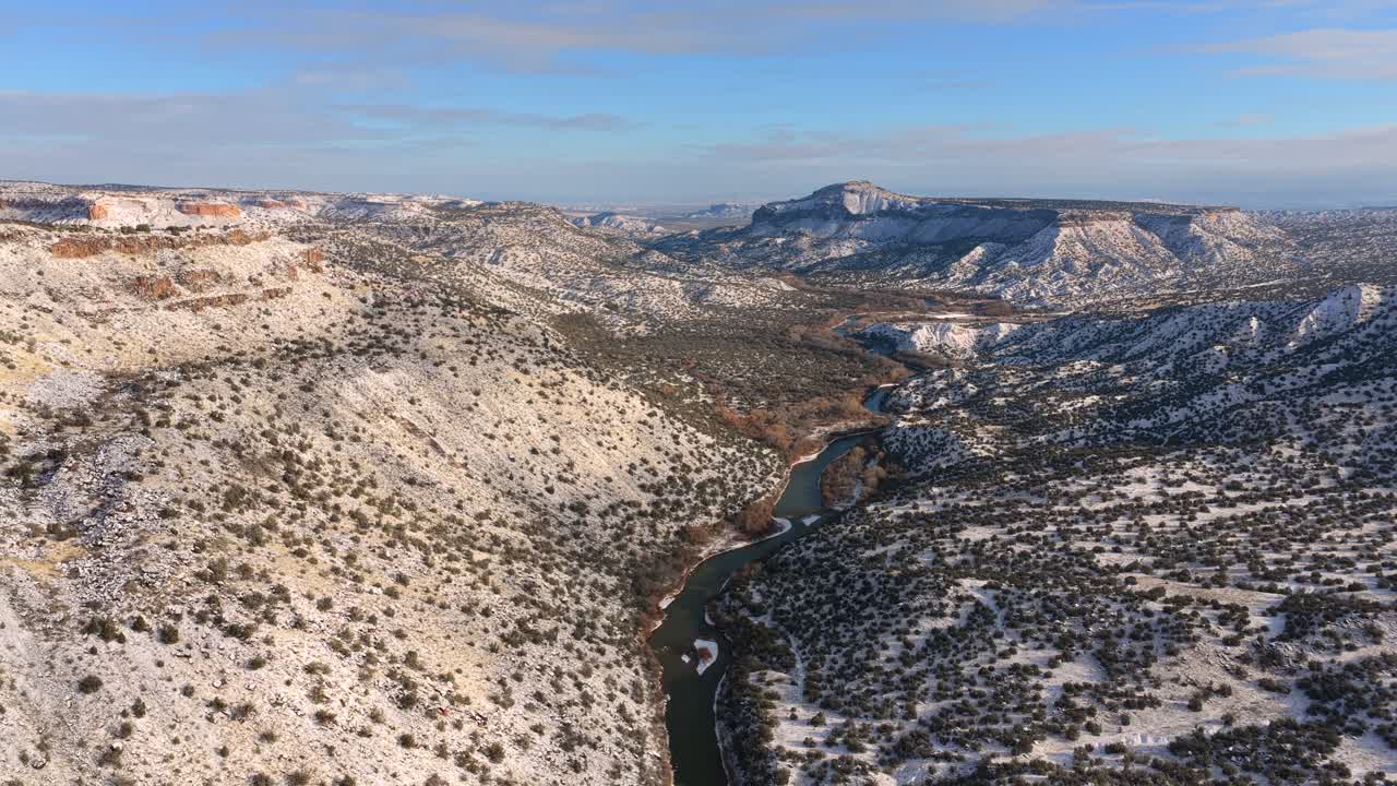 Una vista aerea cinematografica cattura il tortuoso fiume Rio Grande che serpeggia attraverso il canyon White Rock ricoperto di neve al tramonto. Questa vasta ripresa di viaggio mostra le aspre mesas vicino a Los Alamos