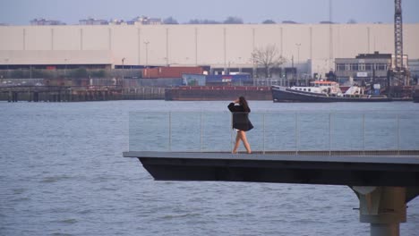 young female in a distance is walking towards the edge of a platform build on water to relax by the river wearing black jacket and shorts near to docks industrial environment windy weather slow motion