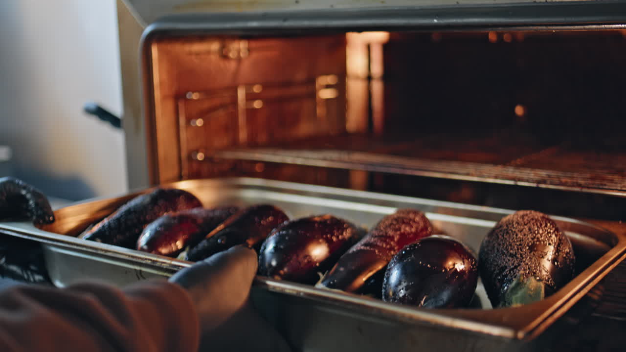Primer plano de un cocinero en la cocina preparando berenjenas y colocando una bandeja en el horno para asarlas