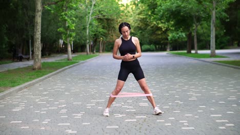 Front-view-of-stylish-woman-makes-side-steps-with-pink-rubber-band-on-legs-on-pavement,-view-on-green-city-park-on-background