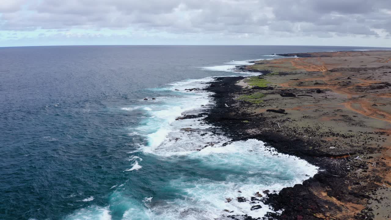 Drone view of rugged volcanic coastline blending into turquoise ocean waves along Mahana Beach, Hawaii. Striking contrast of lava rock, surf, and overcast sky.