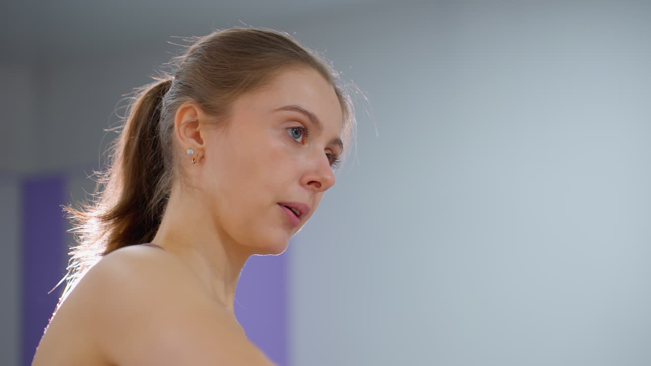 mujer con ojos azules y cabello atado hacia atrás se centra mientras ayuda con un estiramiento del brazo, el fondo presenta un entorno de gimnasio borroso con iluminación suave