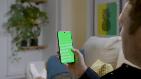 Man-sitting-on-sofa-using-cell-phone-browsing-social-media,-device-screen-in-green-chroma-for-graphic-insertion