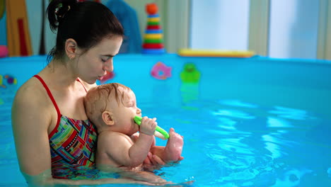 woman with a little baby boy in the swimming pool. adorable infant holds the toy in his mouth. side view.