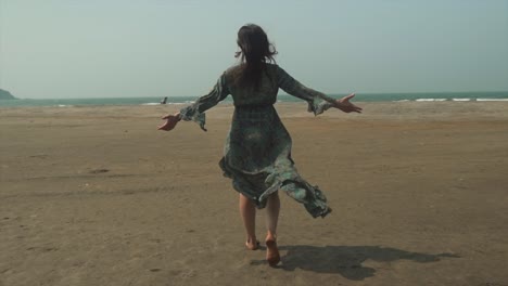 niña con los brazos afuera caminando descalza a través de la brisa con el viento que fluye más allá de su vestido largo en la playa