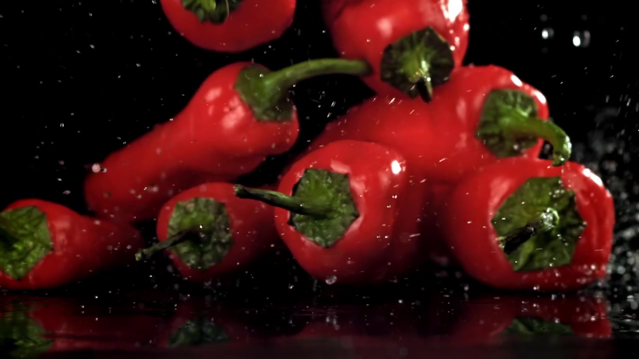 Červené chilli papričky padající a stříkající na mokrý černý povrch ve zpomaleném záběru.