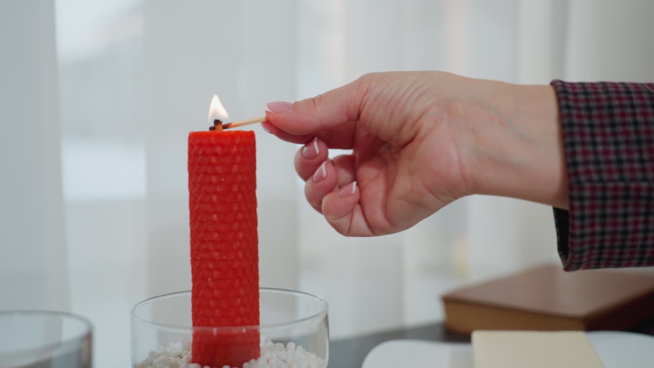 primer plano de mano elegante encendiendo una vela roja con cerilla, creando un ambiente cálido, candelabro de vidrio con cuentas decorativas, mantequilla en el plato y libro en el fondo