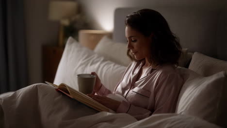 woman reading a book and drinking from a mug in bed at night