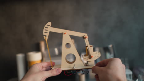 pov video of hands raising a working wooden oil pumpjack model with motor at eye level on a desk for inspection