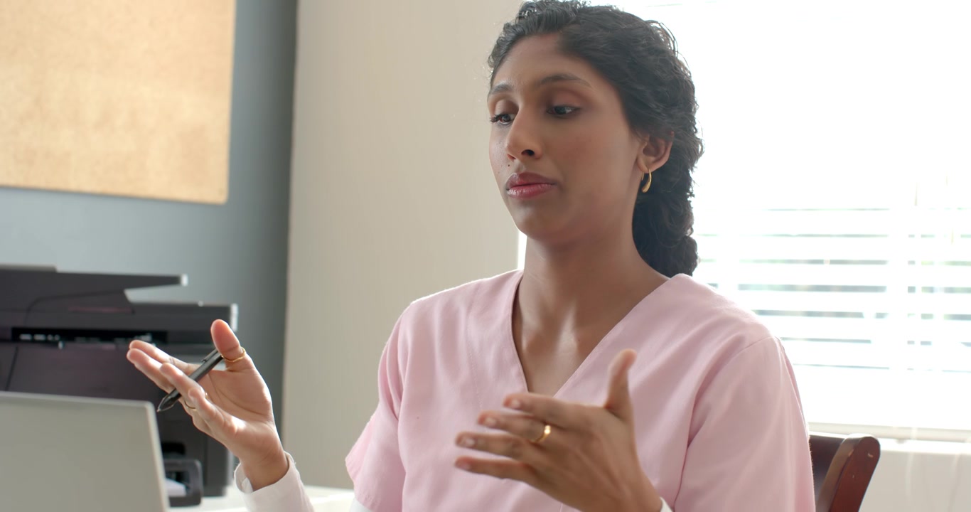 Woman medic in pink scrubs typing, pausing, speaking, explaining chart on laptop with pen at desk