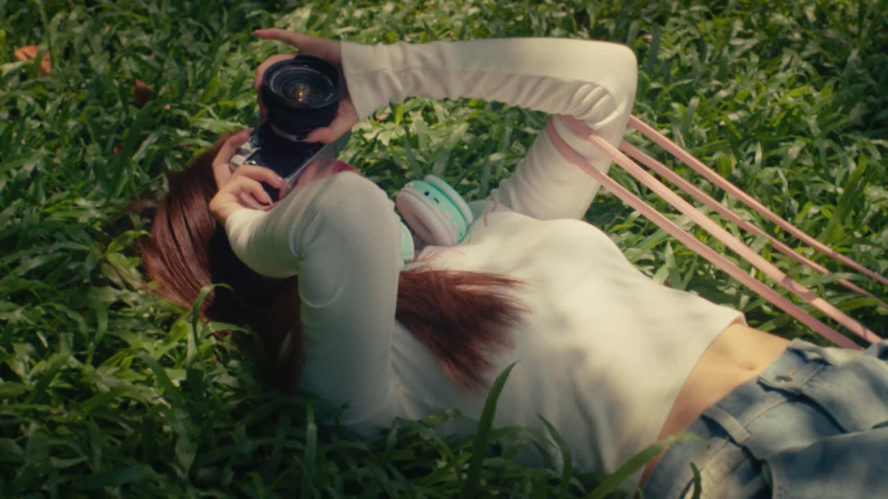 여성 모델이 공원의 초록 잔디에 누워있는 동안 그녀의  셔츠에 나무 잎의 전문 카메라 그림자를 찍고 있습니다.