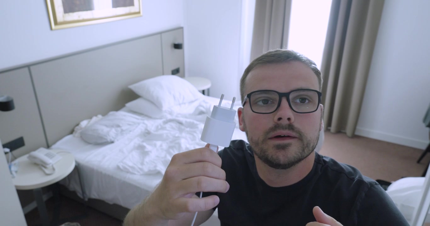Uomo che tiene un adattatore di corrente e lo indica in una camera d'albergo, evidenziando il problema della scarsità di prese elettriche. Concetto di viaggio, ricarica di dispositivi e comodità dell'alloggio