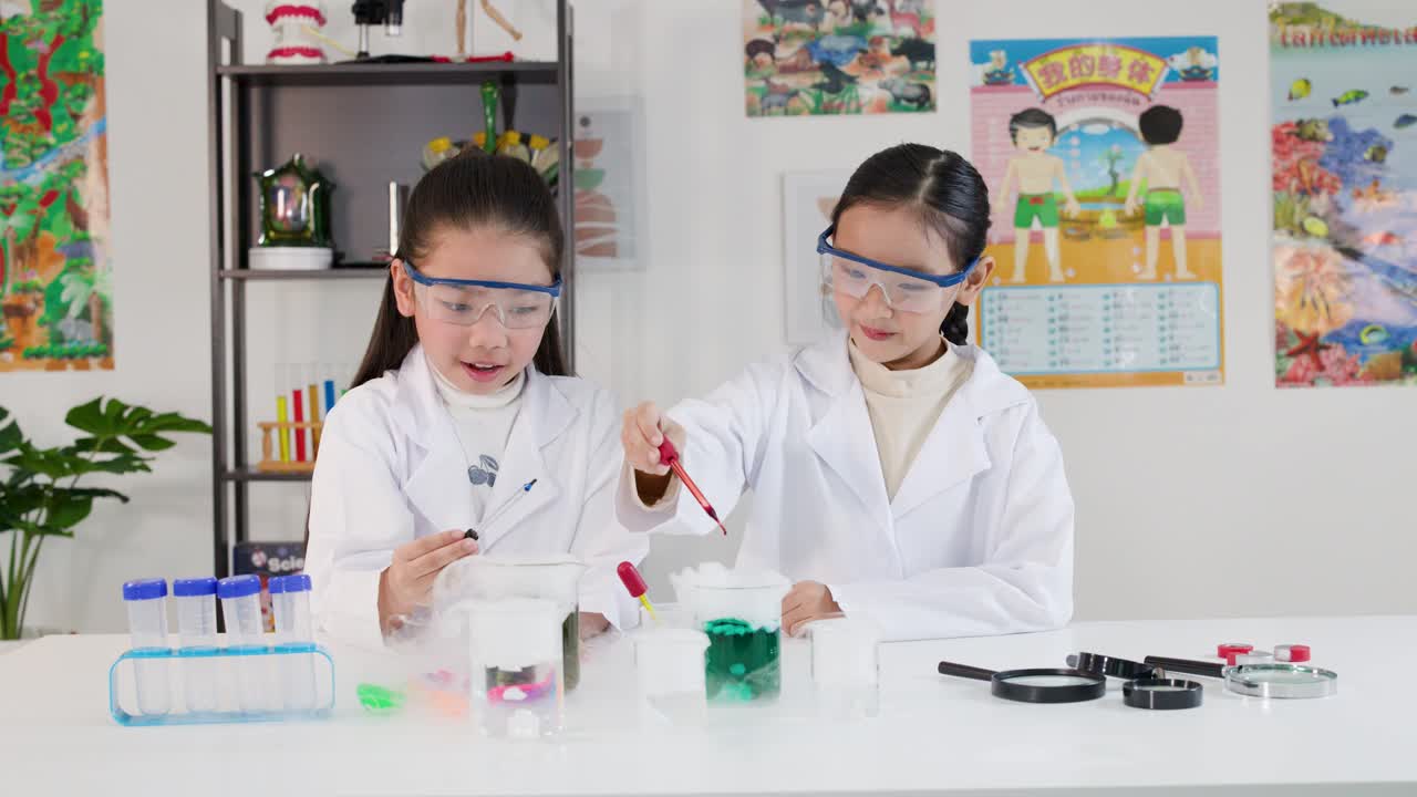 Aasialaiset lapset laboratoriovaatteissa ja suojalaseissa käyttävät pipettejä kemian kokeissa luokkahuoneessa.