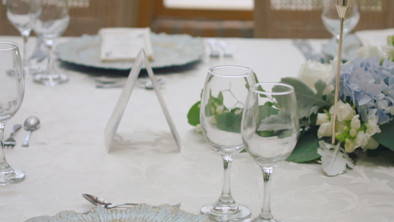 A detailed clip of an elegantly decorated wedding table. It features stemware, a white tablecloth, plates, cutlery, cloth napkins, a delicate floral arrangement, and a personalized invitation.