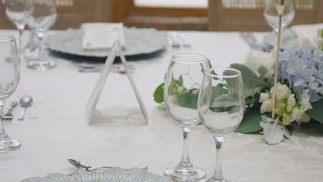 a detailed clip of an elegantly decorated wedding table. it features stemware, a white tablecloth, plates, cutlery, cloth napkins, a delicate floral arrangement, and a personalized invitation.