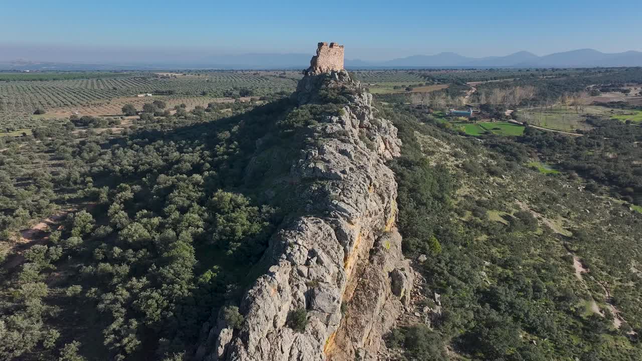 70mm drone flight with a side view of Dos Hermanas Castle in Navahermosa, Toledo. It shows the ruins atop the rocky outcrop, surrounded by olive trees and distant mountains that enhance the landscape
