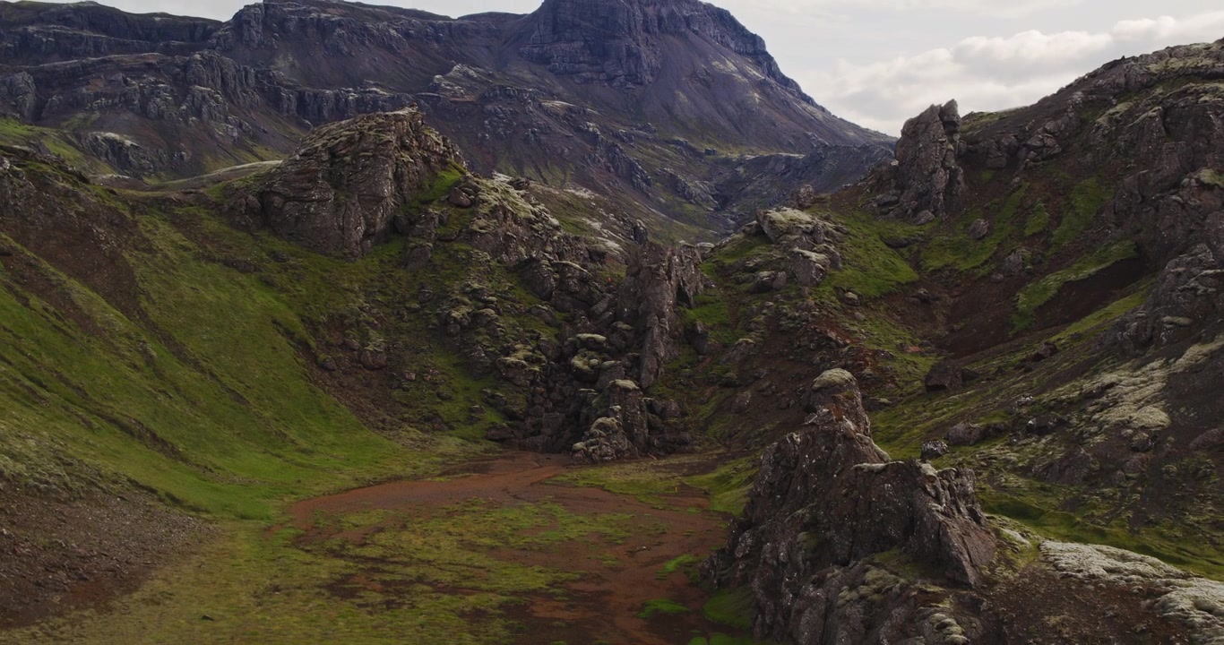 Ripresa aerea con drone in allontanamento con un bell'effetto parallasse di un sentiero escursionistico nelle montagne dell'Islanda. Un piccolo escursionista con una maglia rossa brillante cammina al centro dell'inquadratura, a dimostrazione della vastità del paesaggio.