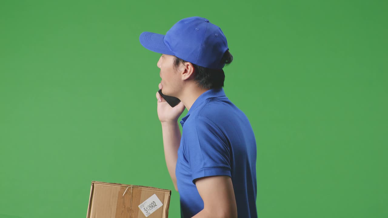 vista lateral de cerca de un mensajero masculino asiático en uniforme azul caminando y hablando en teléfono inteligente mientras entrega una caja de cartón en el fondo de la pantalla verde en el estudio