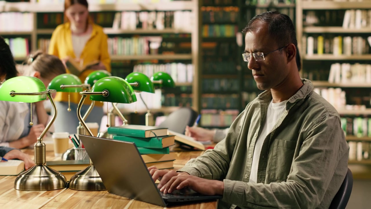 Adult man concentrating on note taking on laptop at wooden library desk