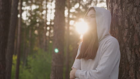 young woman standing in a forest with the sun shining behind her