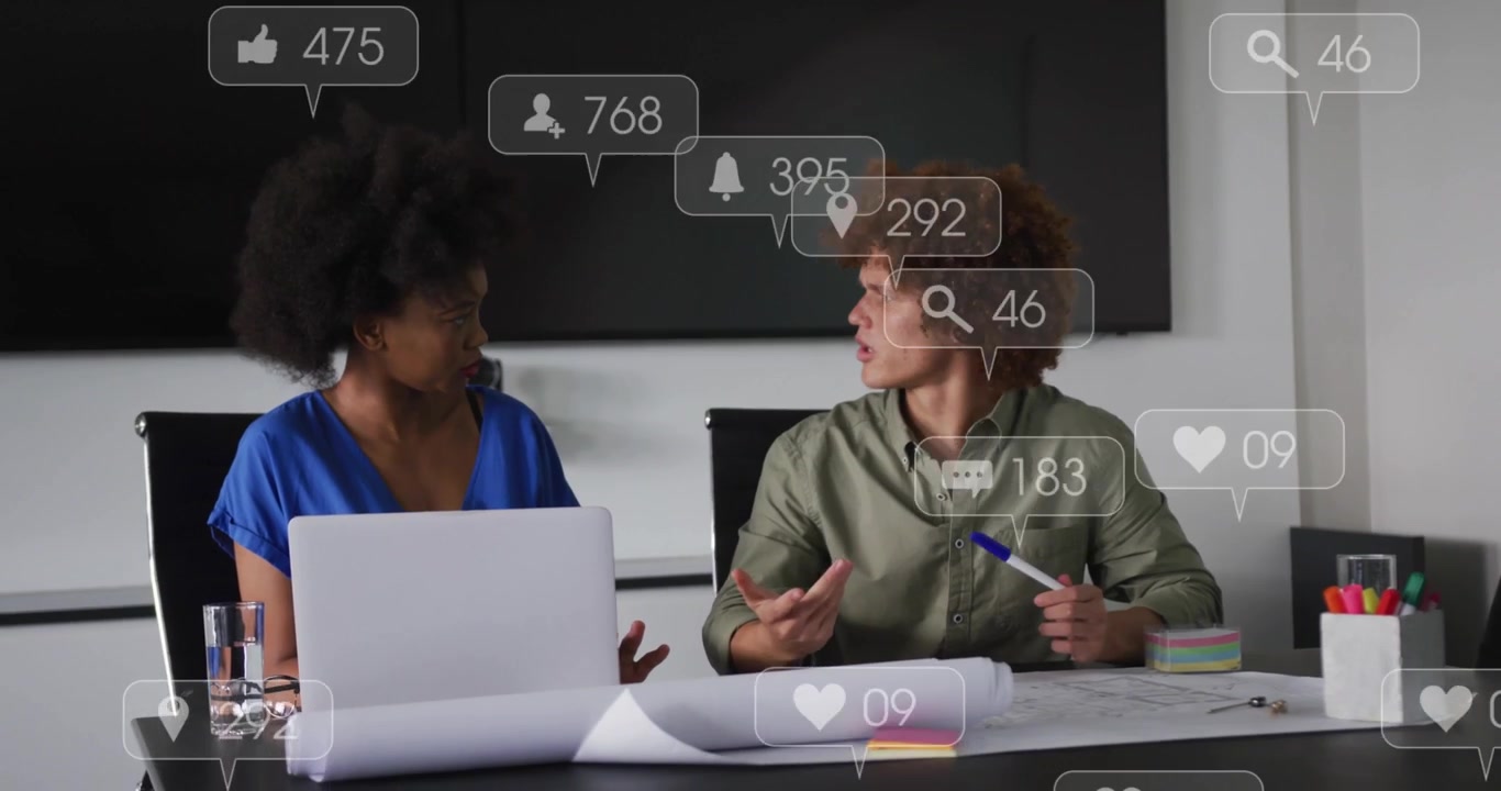 Woman pointing at laptop plans, man holding pen sparking social icons flooding table for marketing