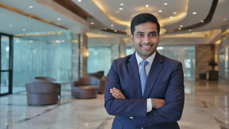 confident indian entrepreneur smiling with arms crossed, standing in elegant corporate building lobby, showcasing leadership and success in business