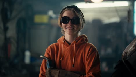 una adolescente rubia con gafas de protección se encuentra en un taller con una sudadera naranja, sosteniendo un