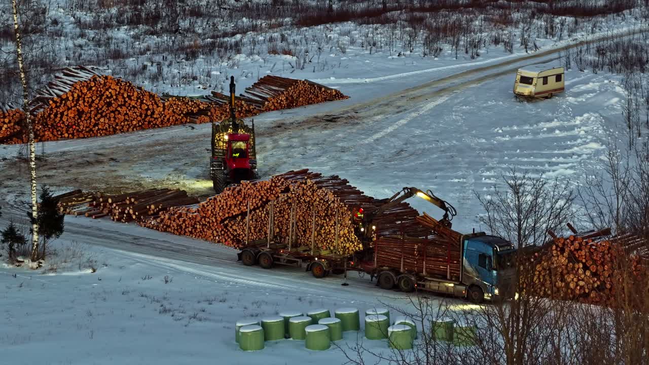 Forestry machinery loading logs onto a truck for transport across a snow-covered road, aerial view