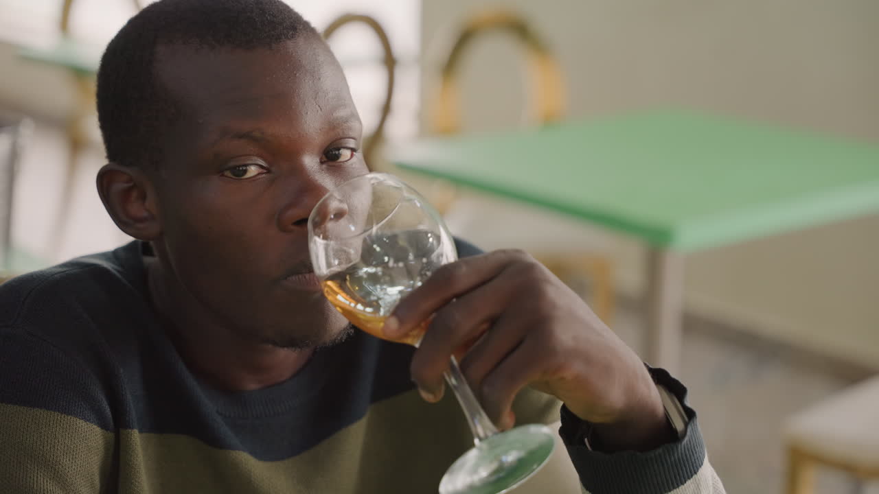 Homme noir pensif après une gorgée, regardant au loin, pause contemplative et tension silencieuse, légère inquiétude et prise de décision, pull décontracté et verre vide, ambiance de bistro en fin de soirée.