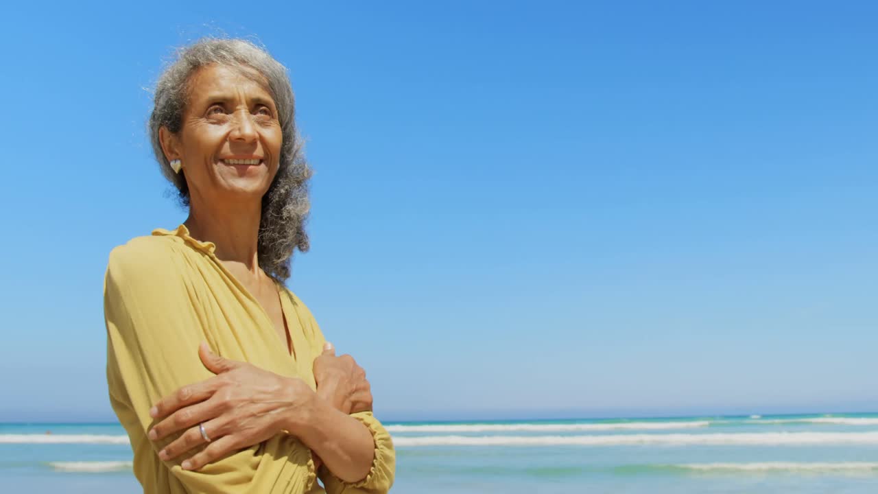 Front view of happy active senior African American woman with arms crossed standing on the beach 4k