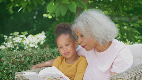 Großmutter-Sitzt-Draußen-Im-Garten-Mit-Enkelin-Zu-Hause-Und-Liest-Gemeinsam-Ein-Buch