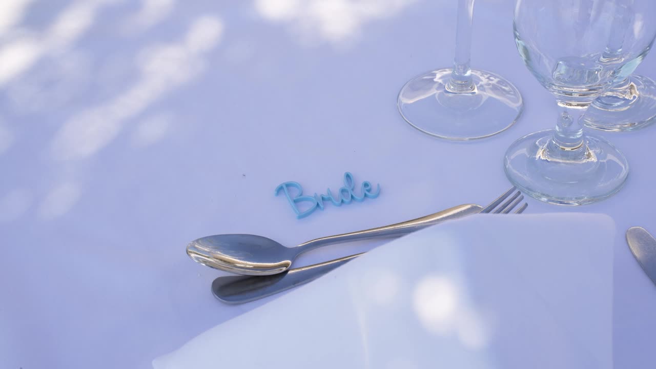 An elegant table is set for a wedding celebration. A name card reading 'Bride' sits beside shiny silverware and stylish glassware under a sunny sky near an outdoor venue