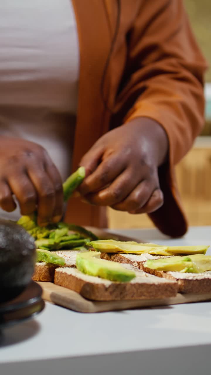 Video verticale: Vlogger culinaria afroamericana che spalma fette di avocado su toast di pane per una scelta salutare.