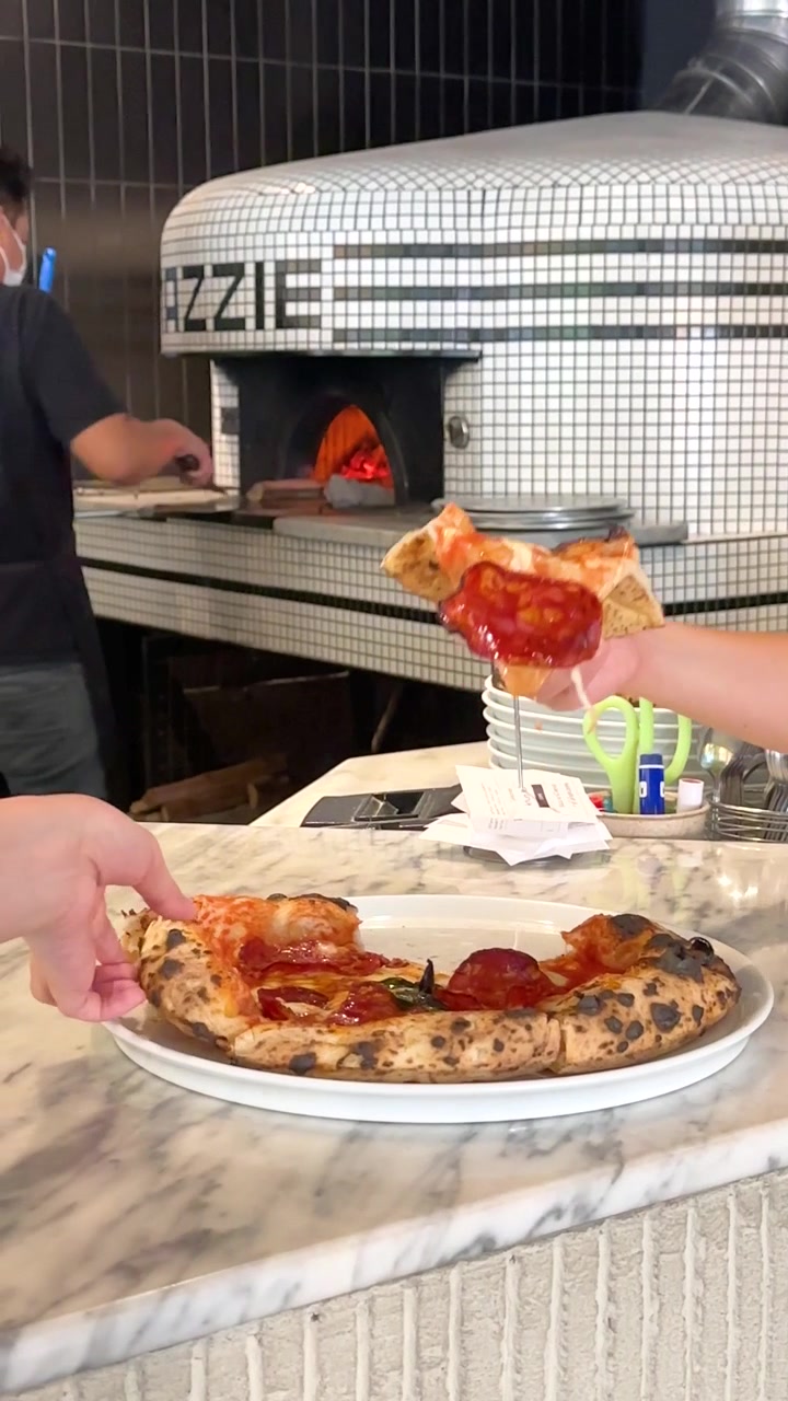 Primo piano di una mano che solleva una fetta di pizza al salame piccante da un bancone di marmo vicino a un forno a legna in mattoni