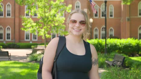 girl student with a backpack wearing sunglasses smiles in front of a university