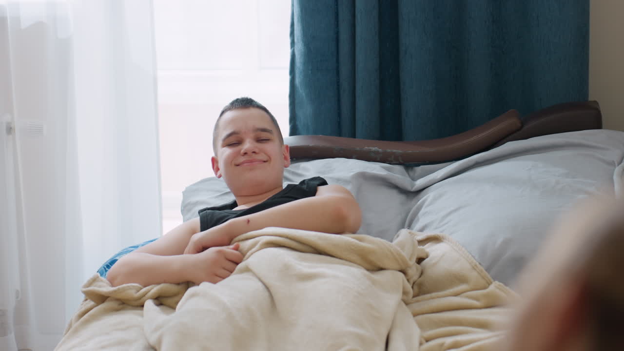 Student wrapped in blanket wakes up stretching in cozy bedroom with soft daylight shining through window while someone nearby watches creating warm family moment of care