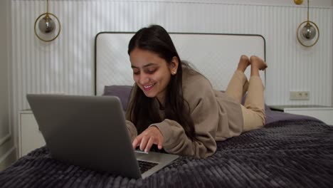 Una-Chica-Morena-Feliz-Con-Un-Suéter-Marrón-Se-Acuesta-En-La-Cama-Y-Escribe-En-Una-Computadora-Portátil-Gris-Mientras-Trabaja-Y-Pasa-Tiempo-En-Línea-En-Un-Apartamento-Moderno-En-El-Dormitorio.