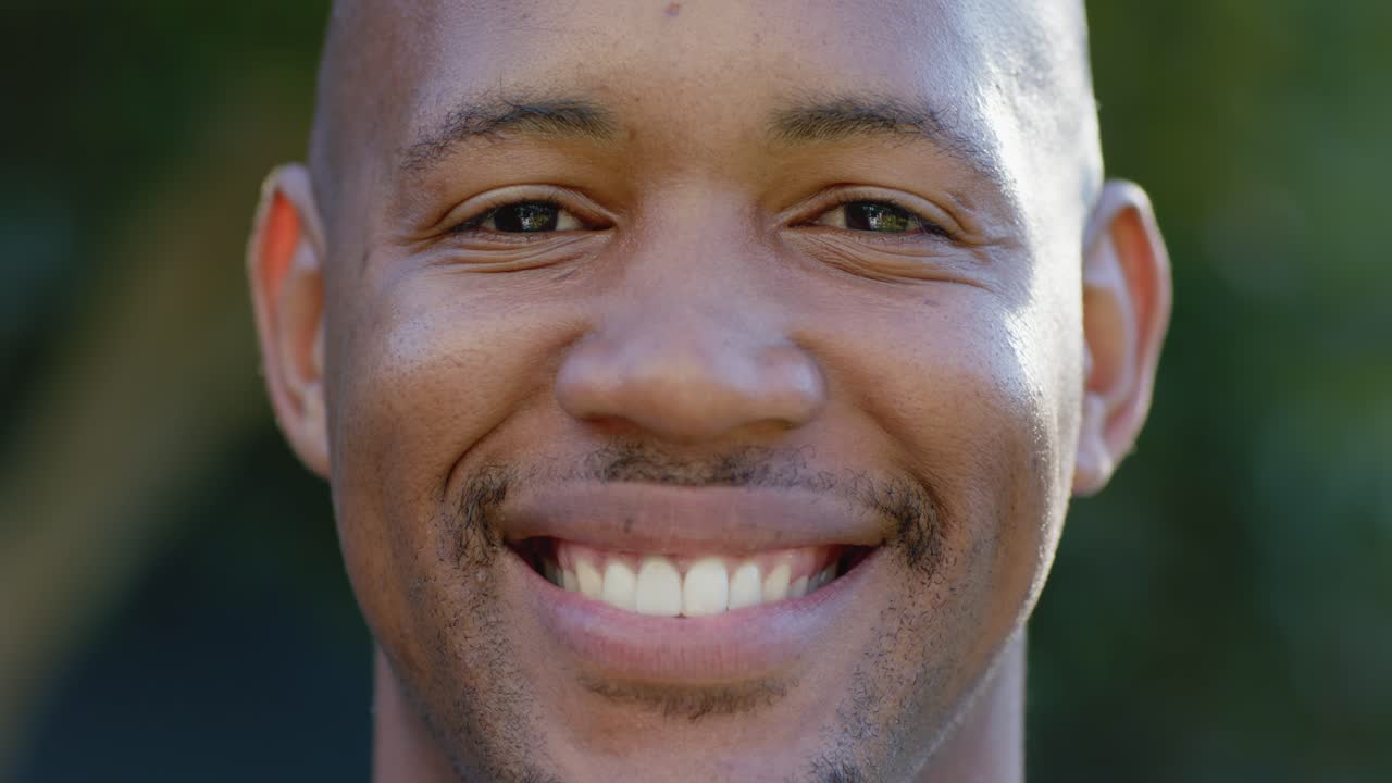 Homem africano adulto de frente para a câmera, sorrindo gradualmente de neutro a um largo sorriso no parque, mostrando alegria