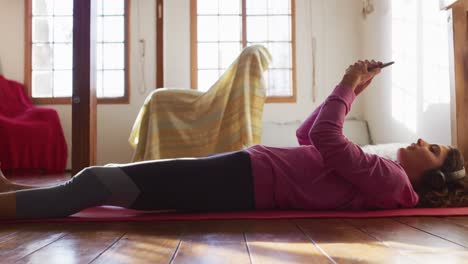 Happy-mixed-race-woman-in-headphones,-lying-on-yoga-mat-using-smartphone-and-smiling-in-living-room