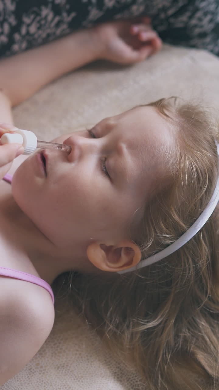 mother gives nose drops to cute sick little girl lying on soft beige bed in light children room close view