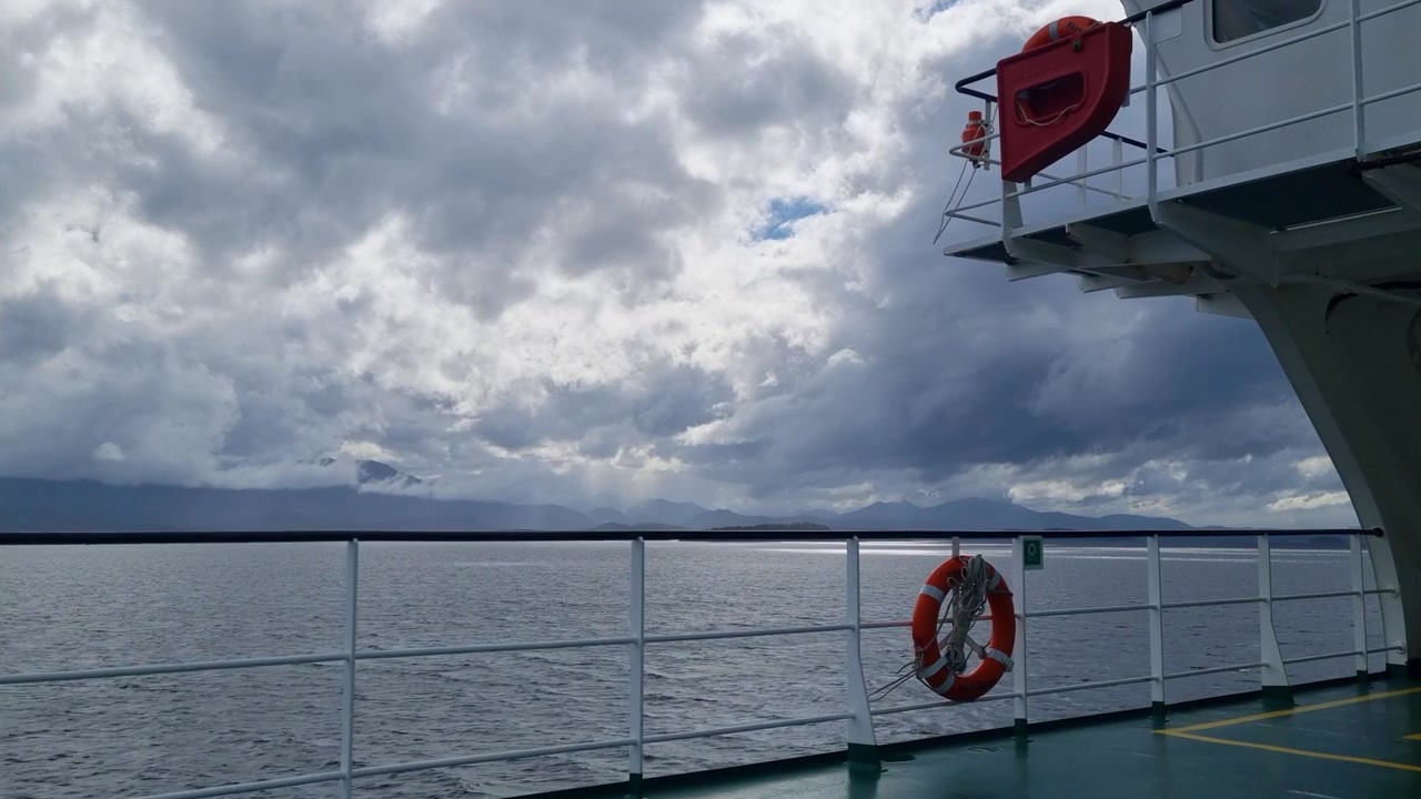 Leeres Fährdeck, das sich durch die Fjorde des chilenischen Patagoniens unter einem stürmischen Himmel bewegt, eine atmosphärische Reise durch wilde Gewässer mit dem Gefühl eines aufziehenden Sturms und eines abgelegenen Abenteuers einfangend.