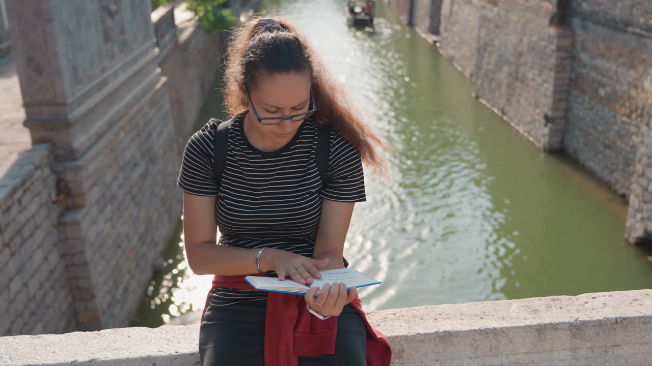 Jovem mulher lendo um mapa em uma ponte de pedra com vista para um canal, verificando a rota com óculos e mochila, luz do sol na alvenaria texturizada e um barco lento ao fundo, clima exploratório tranquilo.