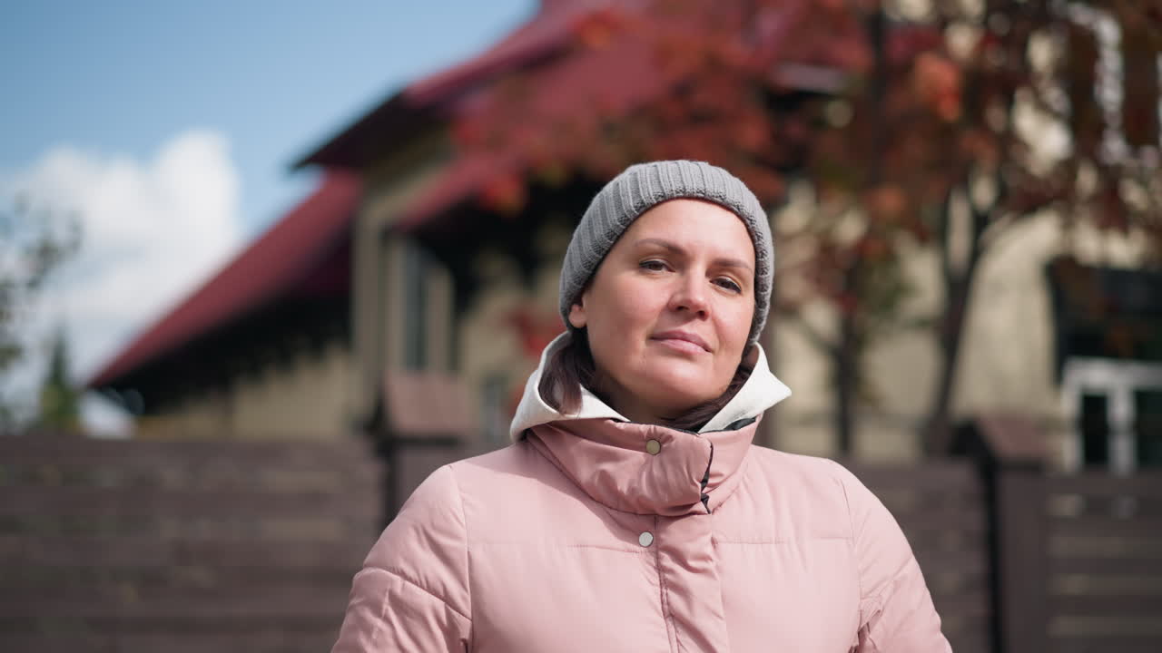 mujer segura en chaqueta rosa y gorra gris sonriendo suavemente con la cabeza inclinada al aire libre, rodeada de árboles borrosos de otoño y telón de fondo de casa vibrante bajo un cielo azul brillante