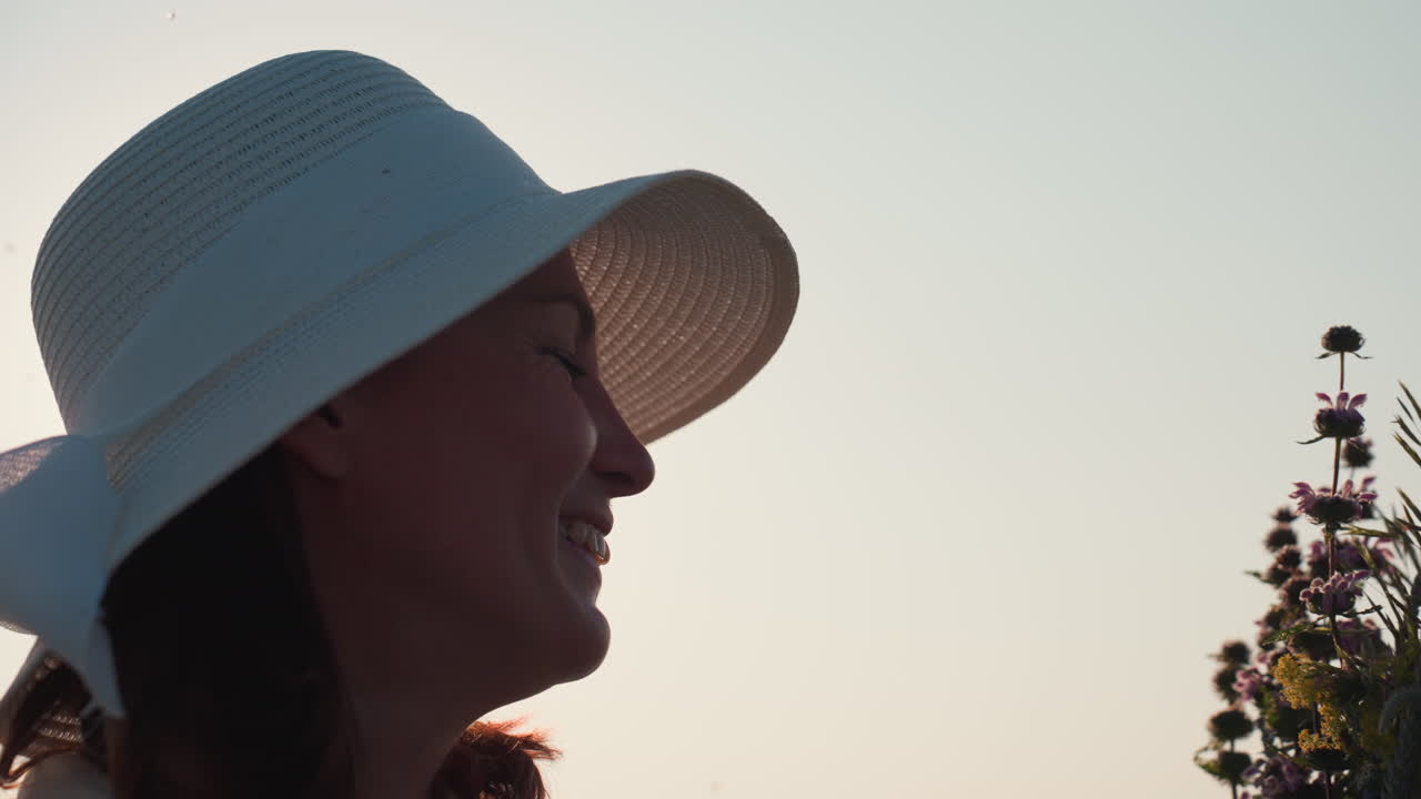 lady in white hat gently lifts wildflowers to nose with serene smile as sun flares behind bouquet, insects drift in soft evening air, capturing tranquil rural moment filled with warmth and peace