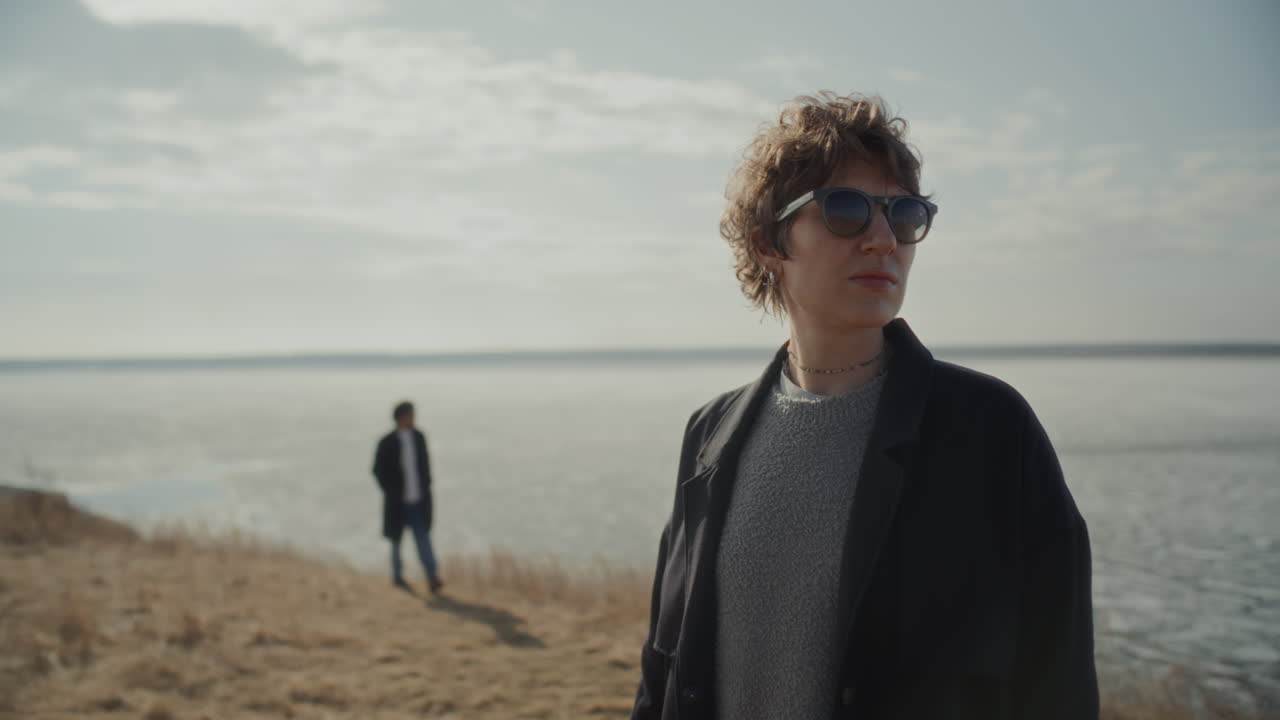 Woman in Sunglasses Standing on Lakeshore with Partner in Background